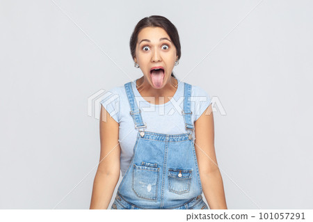 Portrait of disobedient crazy woman wearing denim overalls sticking out tongue and grimacing, aping showing derisive, naughty expression. Indoor studio shot isolated on gray background. 101057291