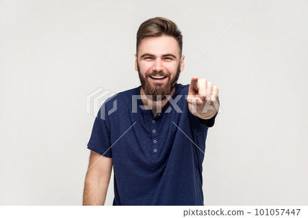 Portrait of amused handsome man with beard wearing dark blue T-shirt laughing out loud, pointing finger to camera, indicating ridiculous idiot. Indoor shot isolated on gray background. Portrait of amused handsome man with beard wearing dark blue T-shirt laughing out loud, pointing finger to camera, indicating ridiculous idiot. Indoor shot isolated on gray background. 101057447