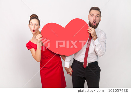 Portrait of amazed shocked man in white shirt and woman in red dress standing together, peeping out from big heart, expressing love. Indoor studio shot isolated on gray background. Portrait of amazed shocked man in white shirt and woman in red dress standing together, peeping out from big heart, expressing love. Indoor studio shot isolated on gray background. 101057488