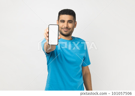 Portrait of cheerful satisfied unshaven man wearing blue T- shirt standing holding out smart phone with empty screen, copy space for advertisement. Indoor studio shot isolated on gray background. 101057506