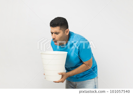 Portrait of sick unhealthy ill unshaven man wearing blue T- shirt standing suffering stomachache, feels nausea and vomits, holding bin in hands. Indoor studio shot isolated on gray background. 101057540