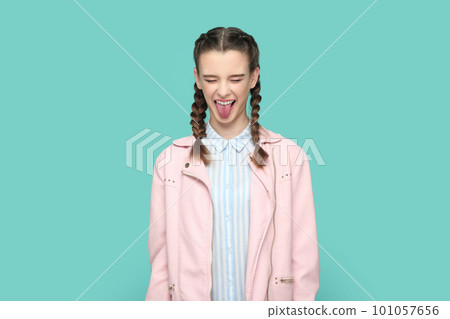 Portrait of funny childish teenager girl with braids wearing pink jacket standing with closed eyes, sticking her tongue out, having fun. Indoor studio shot isolated on green background. Portrait of funny childish teenager girl with braids wearing pink jacket standing with closed eyes, sticking her tongue out, having fun. Indoor studio shot isolated on green background. 101057656