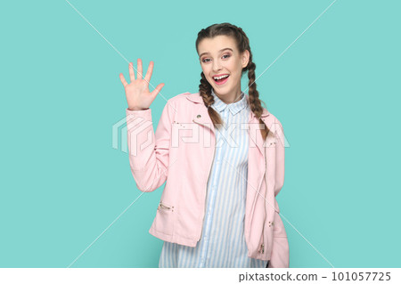 Portrait of friendly lovely teenager girl with braids wearing pink jacket waving hand, saying hello or bye, looking at camera with happiness. Indoor studio shot isolated on green background. 101057725
