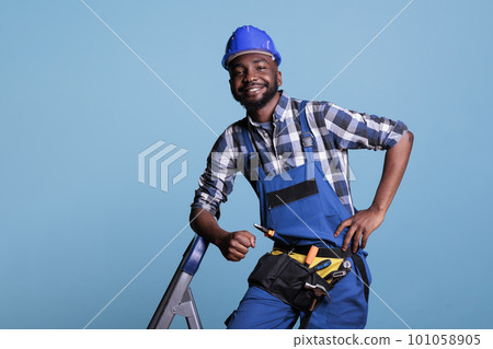Optimistic builder looking at camera with big smile while resting after finishing work in studio shot against blue background. Contractor using tool belt needed for renovation construction tasks. Optimistic builder looking at camera with big smile while resting after finishing work in studio shot against blue background. Contractor using tool belt needed for renovation construction tasks. 101058905
