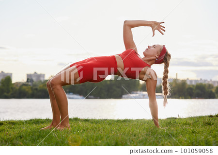 A woman is engaged in animal flow gymnastics, close-up outdoors near the river on green grass in red clothes. A woman is engaged in animal flow gymnastics, close-up outdoors near the river on green grass in red clothes. 101059058