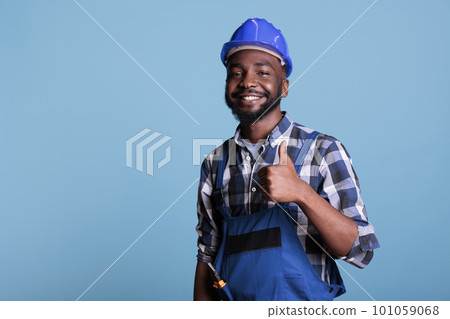 Proud african american professional electrician wearing coveralls and protective helmet. Showing thumb up feeling happy excited about his work while smiling against blue background. background. 101059068