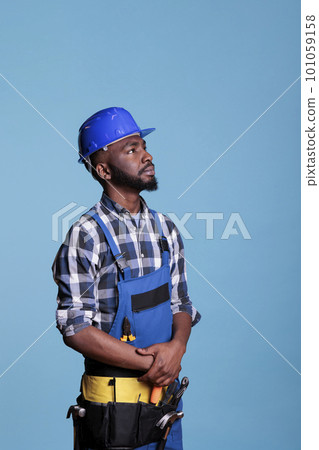 African american builder looking intently up, detailing the tools and materials needed to do the job. Construction worker wearing coveralls and hard hat in studio shot against blue background. 101059158