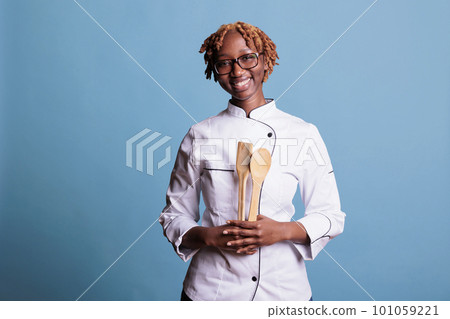 Optimistic african american female chef holding kitchen utensils in studio shot against blue background. Professional female cook at workplace posing smiling for camera. 101059221
