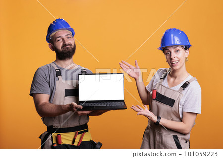 Happy contractors pointing at white laptop display, showing isolated blank screen on wireless pc. Team of builders holding computer with empty copyspace template, posing in studio. 101059223