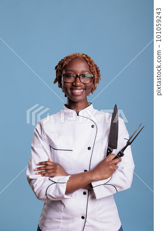 African american female cooker, curly haired posing with professional kitchen knife, sharpener. Smiling afro chef holding kitchen utensils. Portrait of uniformed young female restaurant worker. African american female cooker, curly haired posing with professional kitchen knife, sharpener. Smiling afro chef holding kitchen utensils. Portrait of uniformed young female restaurant worker. 101059243