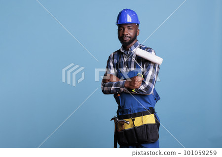 Professional painter looks at camera holding paint roller with overalls and protective helmet, arms crossed over chest. African American construction worker against blue background in studio shot. 101059259