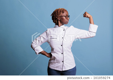 Professional cooking woman flexing arm with pose of power and strength in studio shot. African american chef wearing kitchen uniform looking left arm muscles against blue background. 101059274