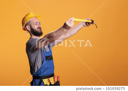 Confident construction worker using tape measure to take measurements and work on refurbishment project. Young male builder holding tapeline and measuring for renovation, wearing tools belt. Confident construction worker using tape measure to take measurements and work on refurbishment project. Young male builder holding tapeline and measuring for renovation, wearing tools belt. 101059275