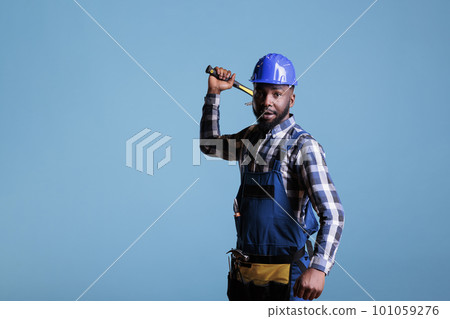 African american construction worker aggressively holding hammer looking at camera. Builder in construction helmet and overalls, looks angry isolated on blue background. African american construction worker aggressively holding hammer looking at camera. Builder in construction helmet and overalls, looks angry isolated on blue background. 101059276