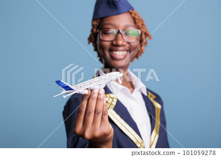 Optimistic african american flight attendant in uniform playing with small custom made airplane toy. Female crew member having some fun before starting a long flight around the world. Optimistic african american flight attendant in uniform playing with small custom made airplane toy. Female crew member having some fun before starting a long flight around the world. 101059278