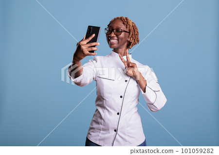 Afro haired female kitchen helper with glasses waving cheerfully, making video conference on mobile phone. Young african american chef taking selfie, self portrait with smartphone. Afro haired female kitchen helper with glasses waving cheerfully, making video conference on mobile phone. Young african american chef taking selfie, self portrait with smartphone. 101059282