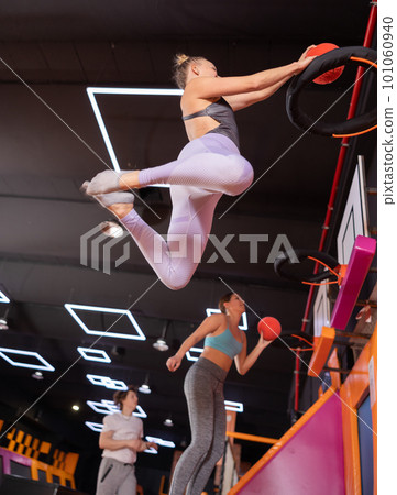 Active European female in sportswear playing basketball while bouncing on colorful trampolines indoors in leisure sports center Active European female in sportswear playing basketball while bouncing on colorful trampolines indoors in leisure sports center 101060940