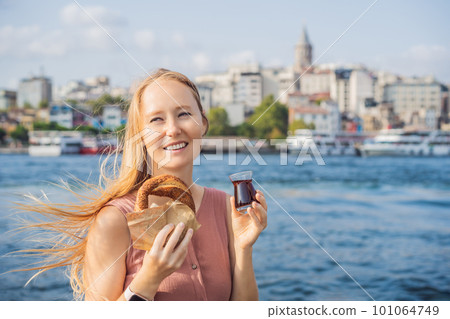 Woman in Istanbul having breakfast with Simit and a glass of Turkish tea. Glass of Turkish tea and bagel Simit against golden horn bay and the Galata Tower in Istanbul, Turkey. Turkiye 101064749