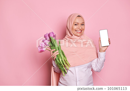 Gorgeous Middle-Eastern Muslim woman in pink hijab, showing at camera a smartphone with white blank digital screen with ad space for mobile application, posing with tulips on isolated pink background 101065113