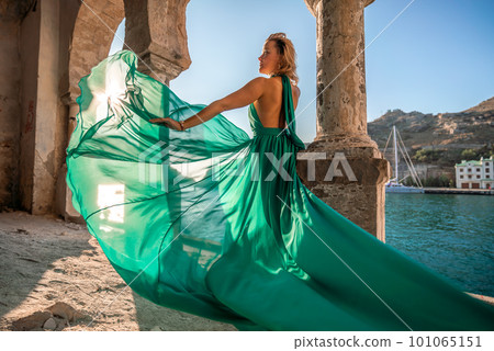 Rear view of a happy blonde woman in a long mint dress posing against the backdrop of the sea in an old building with columns. Girl in nature against the blue sky. Rear view of a happy blonde woman in a long mint dress posing against the backdrop of the sea in an old building with columns. Girl in nature against the blue sky. 101065151