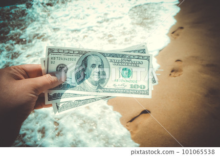 Girl holding money bill of 300 dollars on background of sea ocean waves with white foam and sand wet beach with footprints in sand close-up. Concept finance money holiday traveling dollars vacation Girl holding money bill of 300 dollars on background of sea ocean waves with white foam and sand wet beach with footprints in sand close-up. Concept finance money holiday traveling dollars vacation 101065538