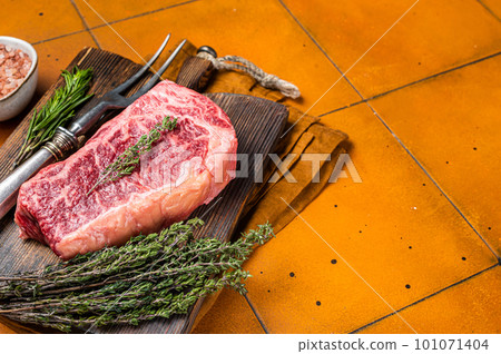 Raw Wagyu striploin, club steak on wooden board with thyme and rosemary ready for cooking. Orange background. Top view. Copy space 101071404