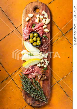 Typical italian antipasto, Cheese and meat plate with Prosciutto ham, Parmesan, Blue cheese, Melon and Olives on wooden board. Orange background. Top view Typical italian antipasto, Cheese and meat plate with Prosciutto ham, Parmesan, Blue cheese, Melon and Olives on wooden board. Orange background. Top view 101071749