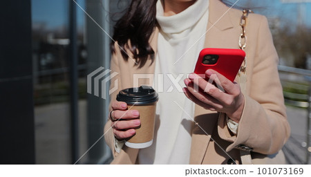 Young happy woman using smartphone and drinking coffee to go outdoors, walking down the city street. 101073169