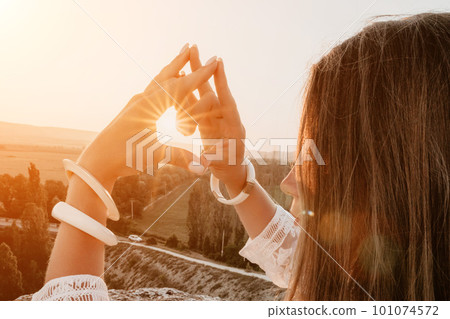 Happy woman in white boho dress making heart sign with hands on sunset in mountains. Romantic woman with long hair standing with her back on the sunset in nature in summer with open hands. Happy woman in white boho dress making heart sign with hands on sunset in mountains. Romantic woman with long hair standing with her back on the sunset in nature in summer with open hands. 101074572