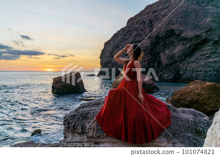 Woman in a red flying dress on the ocean or on the sea beach against the backdrop of the sunset sky. 101074821