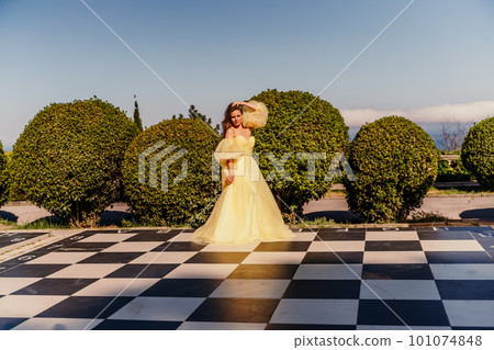 Woman yellow dress chess. A beautiful woman in a long puffy yellow dress poses on a chessboard in the park. 101074848