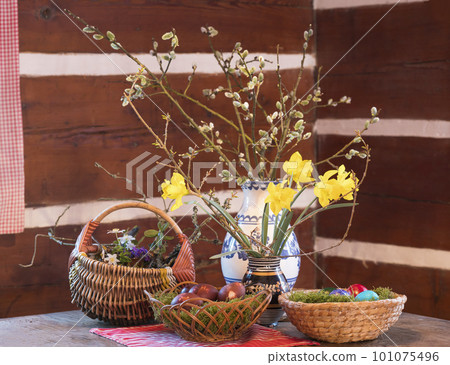 Easter and spring still life with wicker baskets, coloured decorated easter eggs ,moss, feather, vases flowers with narcissus on rustic wooden table in log cabin. Happy Easter holiday card Easter and spring still life with wicker baskets, coloured decorated easter eggs ,moss, feather, vases flowers with narcissus on rustic wooden table in log cabin. Happy Easter holiday card 101075496
