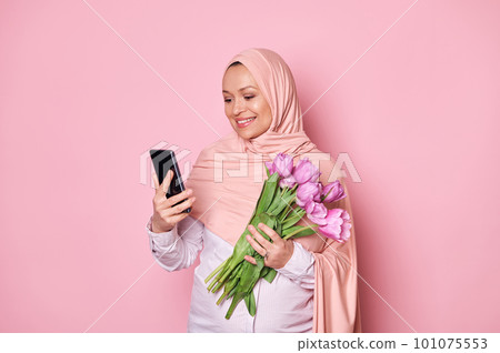 Arab Muslim pregnant woman in pink hijab, holding a bouquet of purple tulips and checking her smartphone, downloading new mobile application, text messaging, smiling on isolated pink color background 101075553