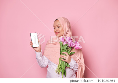 Happy pregnant Arab Muslim woman in pink hijab, with a bouquet of tulips, looking at smartphone with white blank screen, with free advertising space for mobile application, isolated on pink background 101080264