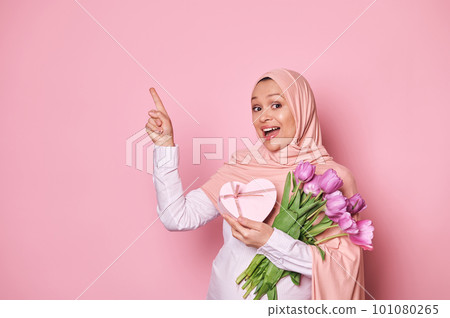 Delighted pregnant Middle-Eastern Muslim woman dressed in elegant pink hijab, with a bouquet of purple tulips and a cute present, pointing at copy advertising space, isolated on pink color background Delighted pregnant Middle-Eastern Muslim woman dressed in elegant pink hijab, with a bouquet of purple tulips and a cute present, pointing at copy advertising space, isolated on pink color background 101080265