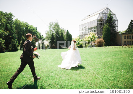 Stylish couple of happy newlyweds. Bride running from groom in the park on their wedding day with bouquet 101081390