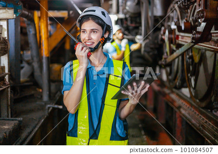 Beautiful woman engineer in factory talking on the radio wearing white helmet and work clothes and hand holding tablet at workplace Beautiful woman engineer in factory talking on the radio wearing white helmet and work clothes and hand holding tablet at workplace 101084507