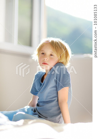 Vertical portrait of cute dreamy schoolboy child in a blue tshirt in the bedroom on sunny day. Preteen boy sitting on the bed and dreaming. 101085133