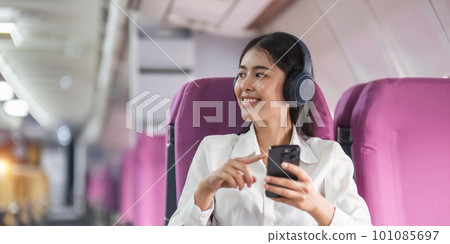 Cheerful female passenger in headphones for noise cancellation watching online movie during intercontinental flight in cabin of aircraft, happy young woman using wifi connection on board 101085697