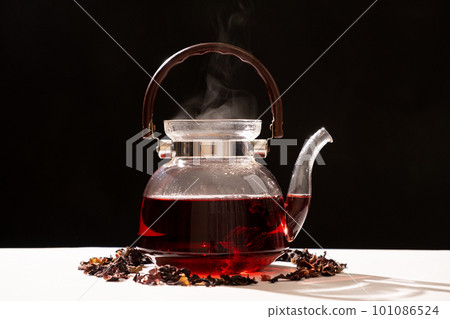 Red hibiscus tea from the petals of a Sudanese rose in a glass teapot, steam from a teapot on a black background Red hibiscus tea from the petals of a Sudanese rose in a glass teapot, steam from a teapot on a black background 101086524