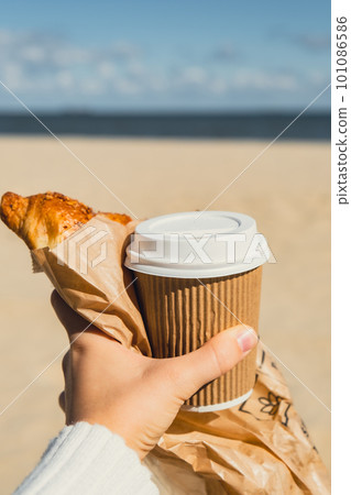 Tasty fresh croissants with coffee in paper cup in female hands on beach sea background. Breakfast outdoors wellbeing concept. FPV. Take away food Tasty fresh croissants with coffee in paper cup in female hands on beach sea background. Breakfast outdoors wellbeing concept. FPV. Take away food 101086586