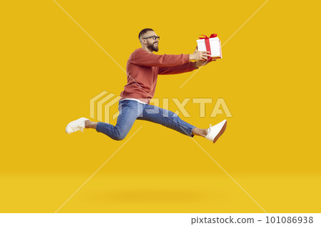 Smiling young man in glasses jumping high holding gift smiling isolated on yellow background. 101086938