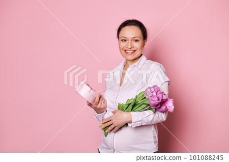 Pregnant middle-aged multi-ethnic woman, smiling looking at camera, expressing positive emotions, getting cute present and bouquet of tulips for Mother's or Women's Day, isolated on pink background Pregnant middle-aged multi-ethnic woman, smiling looking at camera, expressing positive emotions, getting cute present and bouquet of tulips for Mother's or Women's Day, isolated on pink background 101088245