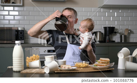 Tired man holding baby and trying to eat while sitting at table in messy kitchen. Single father concept Tired man holding baby and trying to eat while sitting at table in messy kitchen. Single father concept 101088776