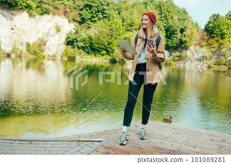 Happy woman tourist with backpack and red hat holding paper map walking in mountains with lake view background. Travel and wanderlust concept. Amazing chill moment.  101089281