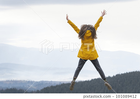 Cheerful girl in bright yellow jacket jumps with hands up to the sky on the mountains background. Travel hiking,tourism 101089412