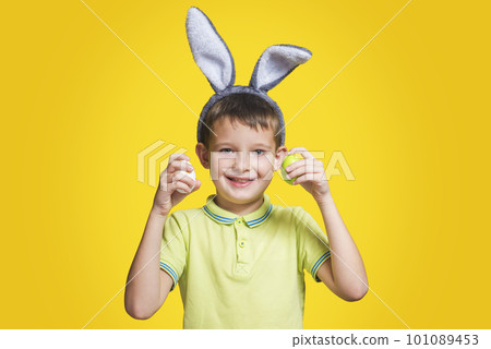 Cute boy with bunny ears holds colored eggs in his hands on yellow background Cute boy with bunny ears holds colored eggs in his hands on yellow background 101089453