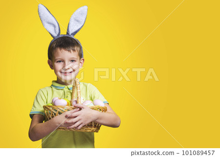 A happy smiling boy wearing Easter bunny ears holds a basket with painted eggs A happy smiling boy wearing Easter bunny ears holds a basket with painted eggs 101089457