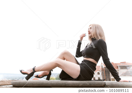 Beautiful slender blonde girl in black clothes wears heels sitting on observation deck and showing off her beautiful legs. Beautiful slender blonde girl in black clothes wears heels sitting on observation deck and showing off her beautiful legs. 101089511