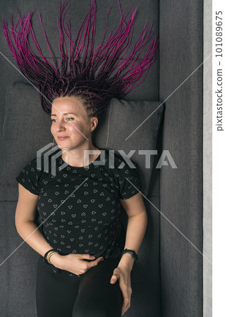Portrait of middle-aged woman with dreadlocks and long hair on gray sofa, looking away. Vertical frame. 101089675